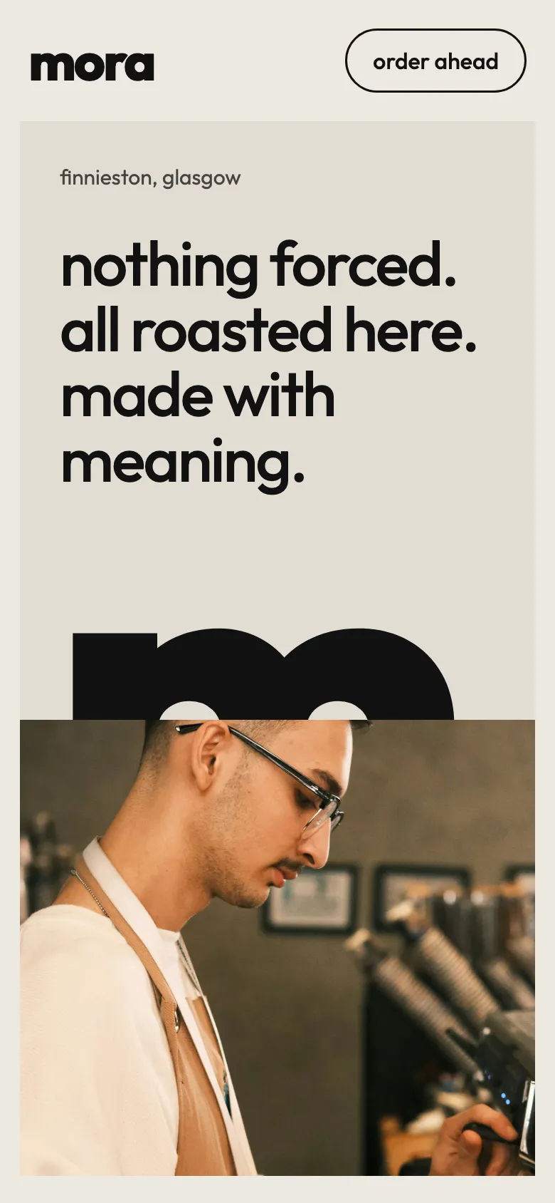 The mora homepage: nothing forced, all roasted here, made with meaning in lowercase type on warm stone, a giant cropped m, a barista in a cream apron at the machine, and opening hours. example site on a phone.
