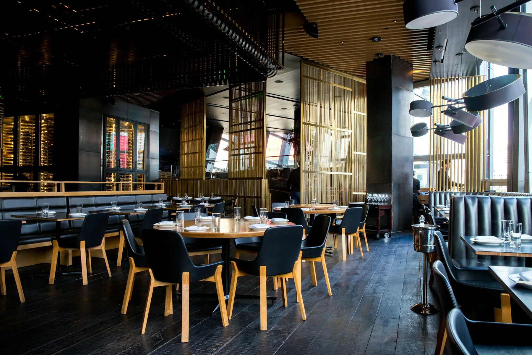 An empty restaurant at night, with dark wood tables, black chairs and low pendant lights.