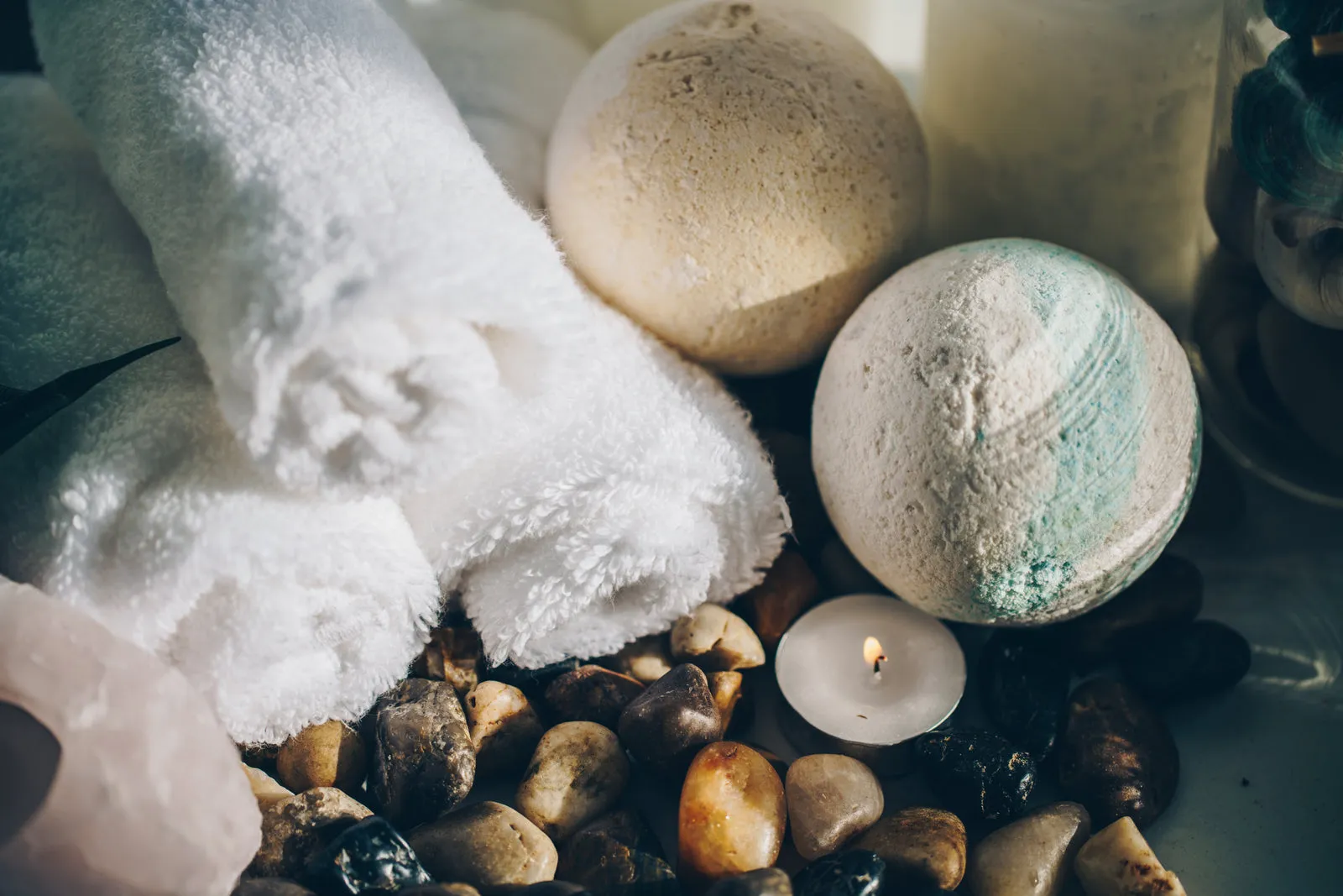 Rolled white towels, bath bombs and a lit candle on pebbles.